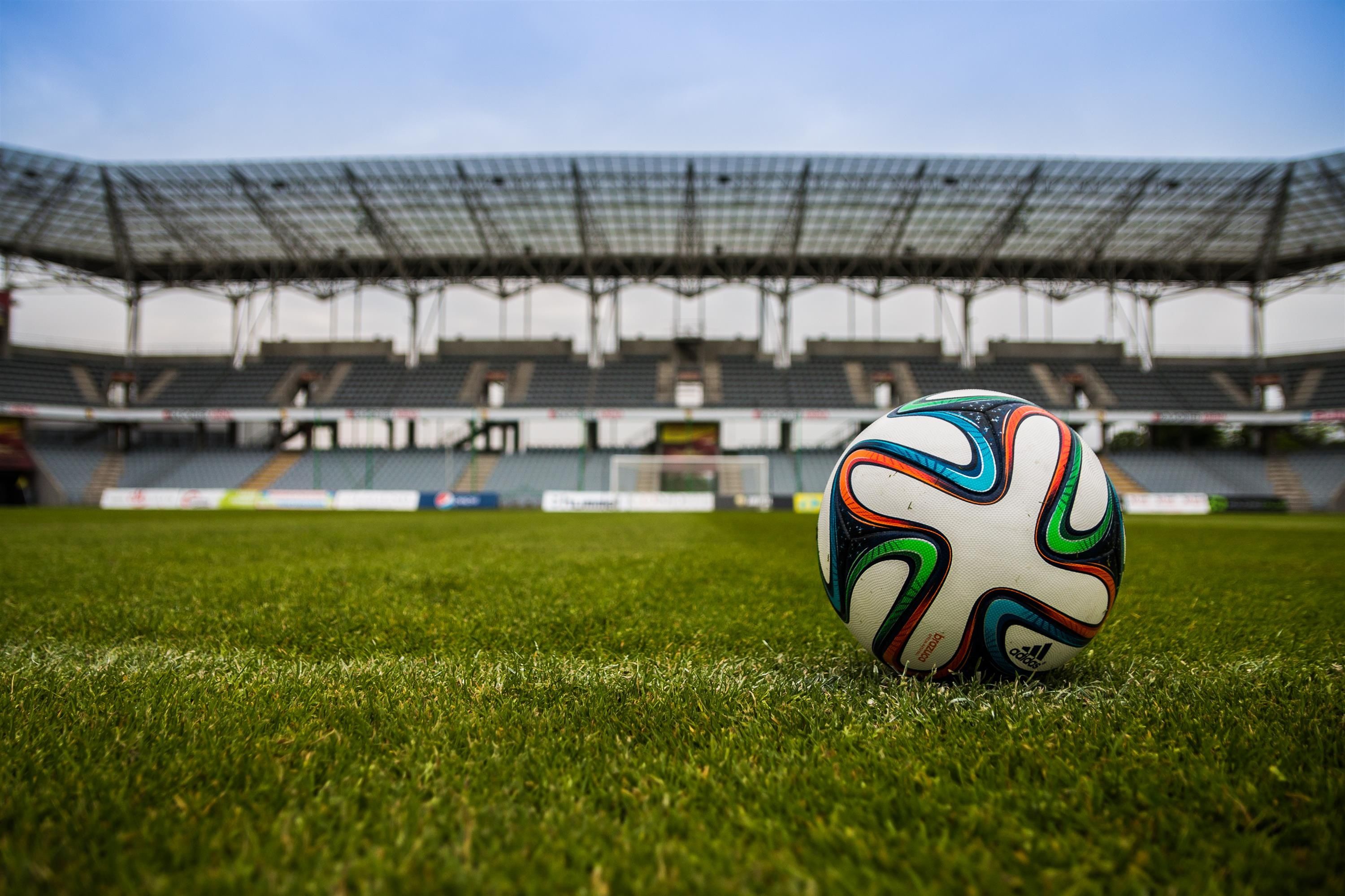 Bola de futebol sobre gramado com arquibancadas desfocadas ao fundo.