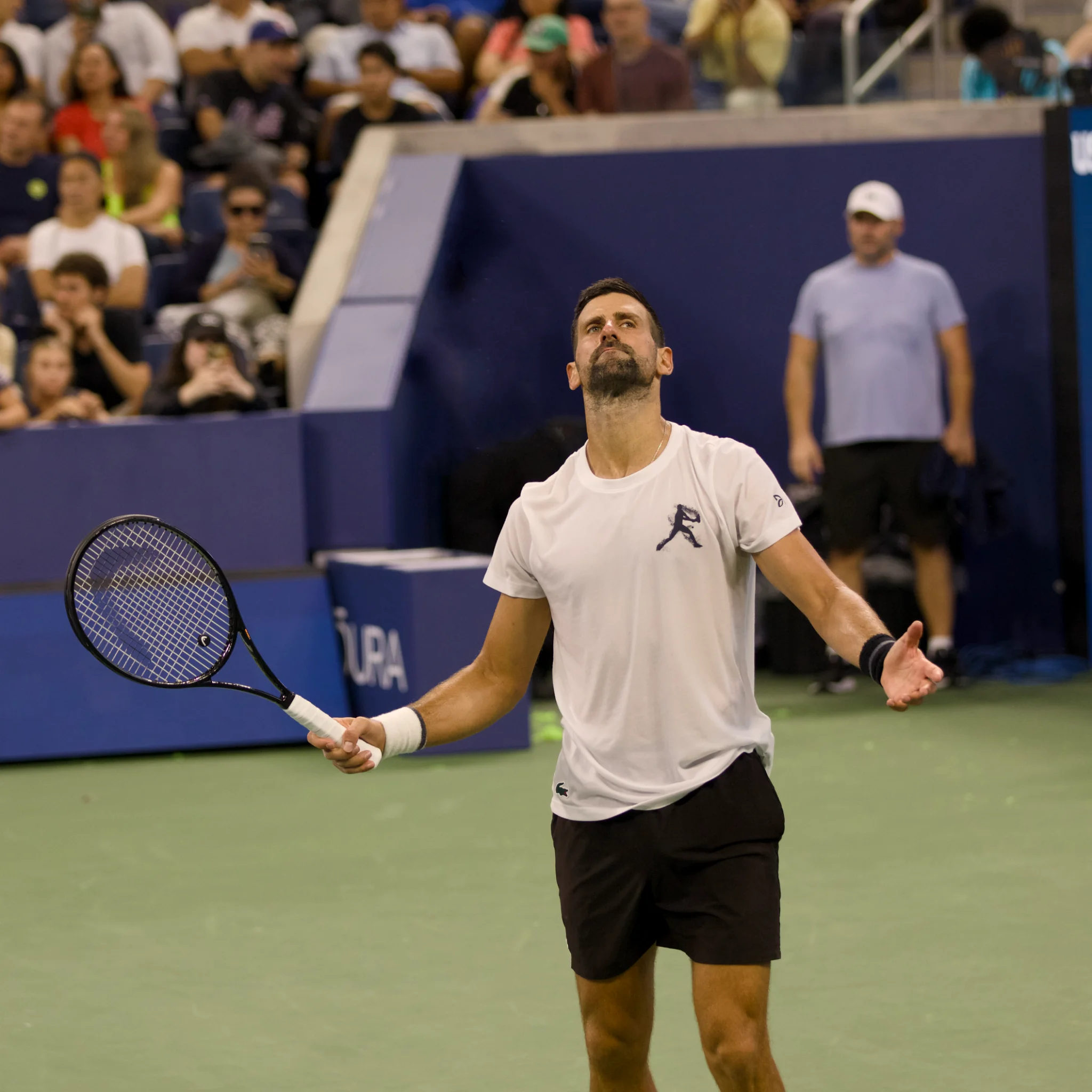Novak Djokovic em quadra com raquete, camiseta branca e shorts pretos.