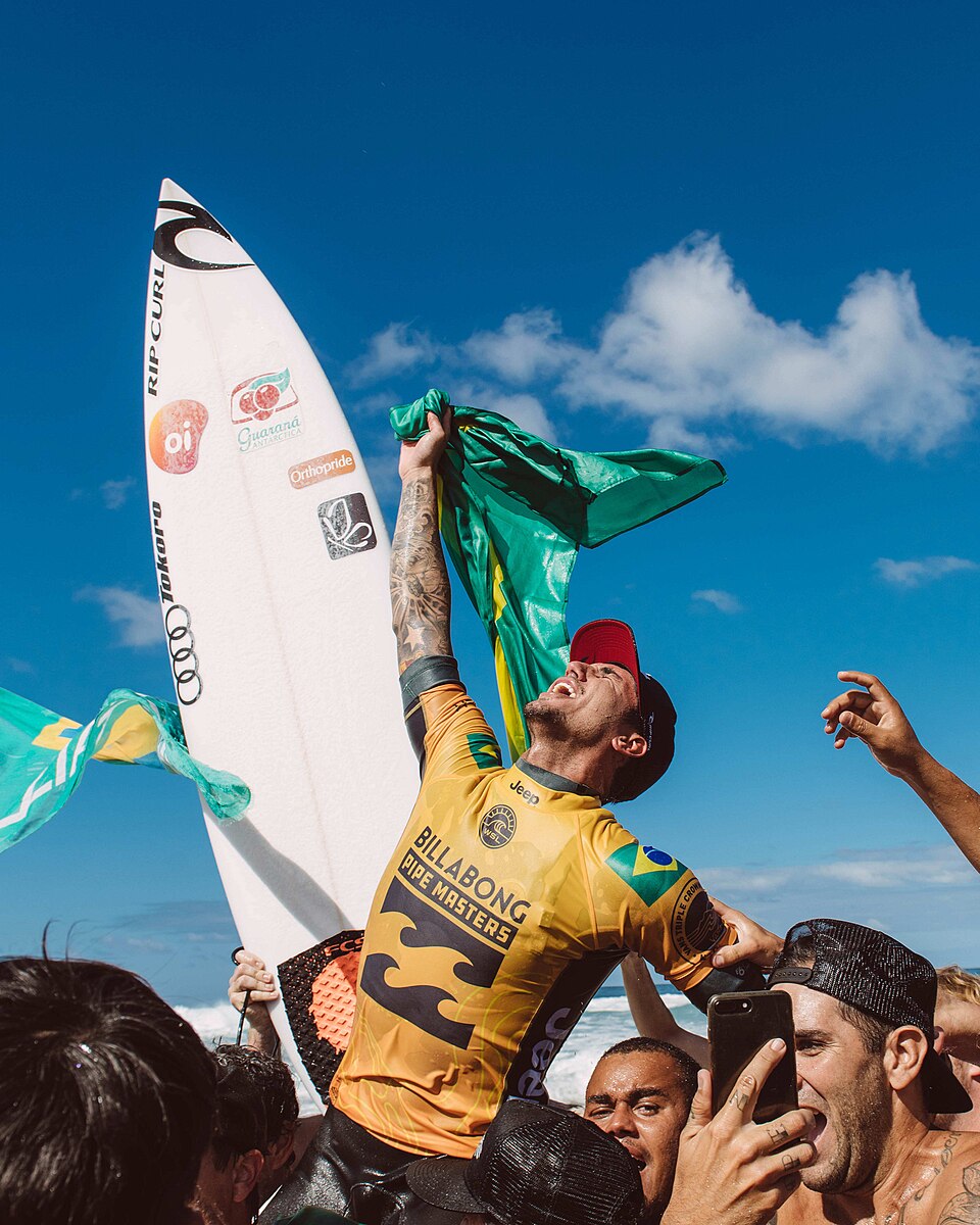 Gabriel Medina de lycra amarela, segurando uma prancha e uma bandeira do Brasil em meio à comemoração.