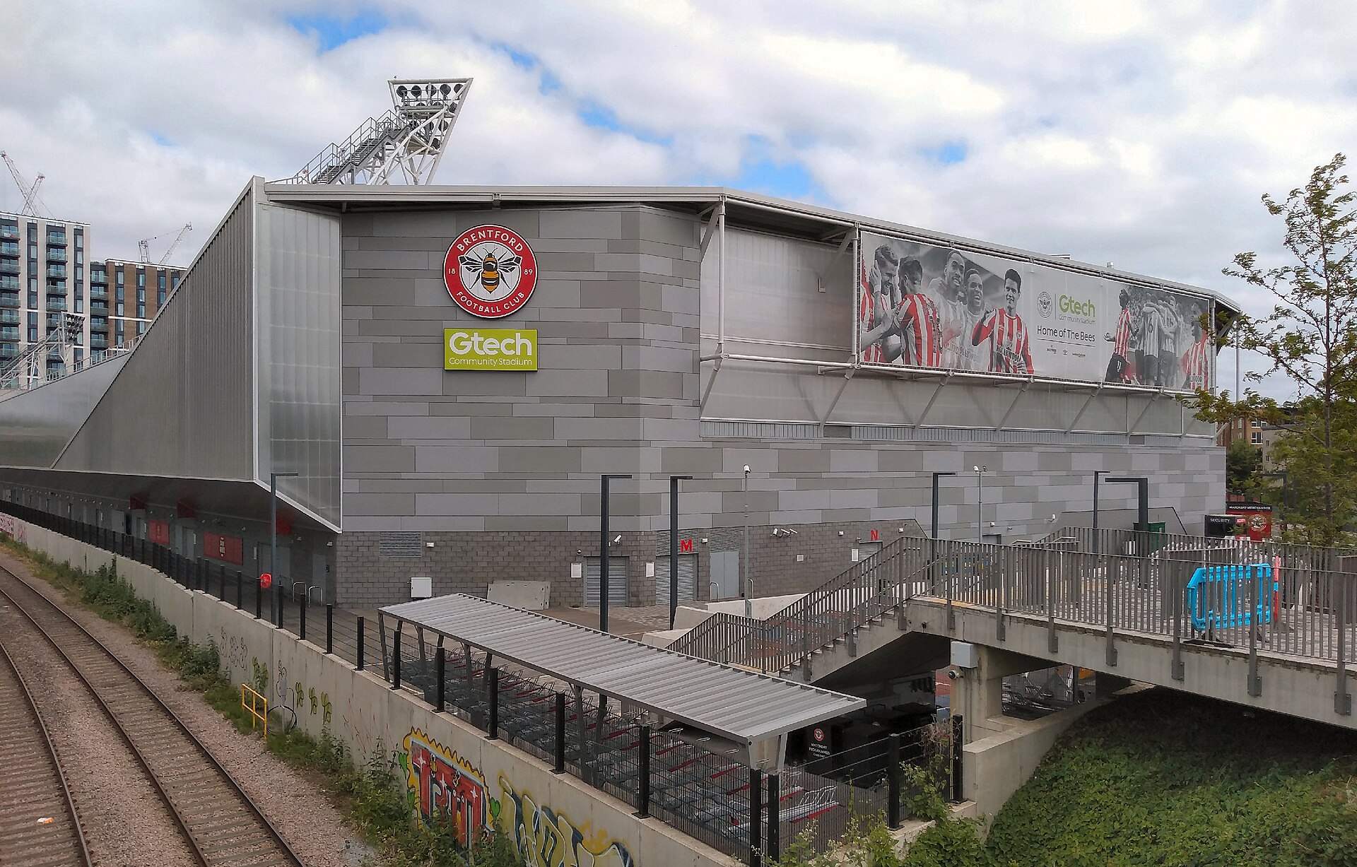 Exterior do Gtech Community Stadium junto à linha férrea e à passarela.