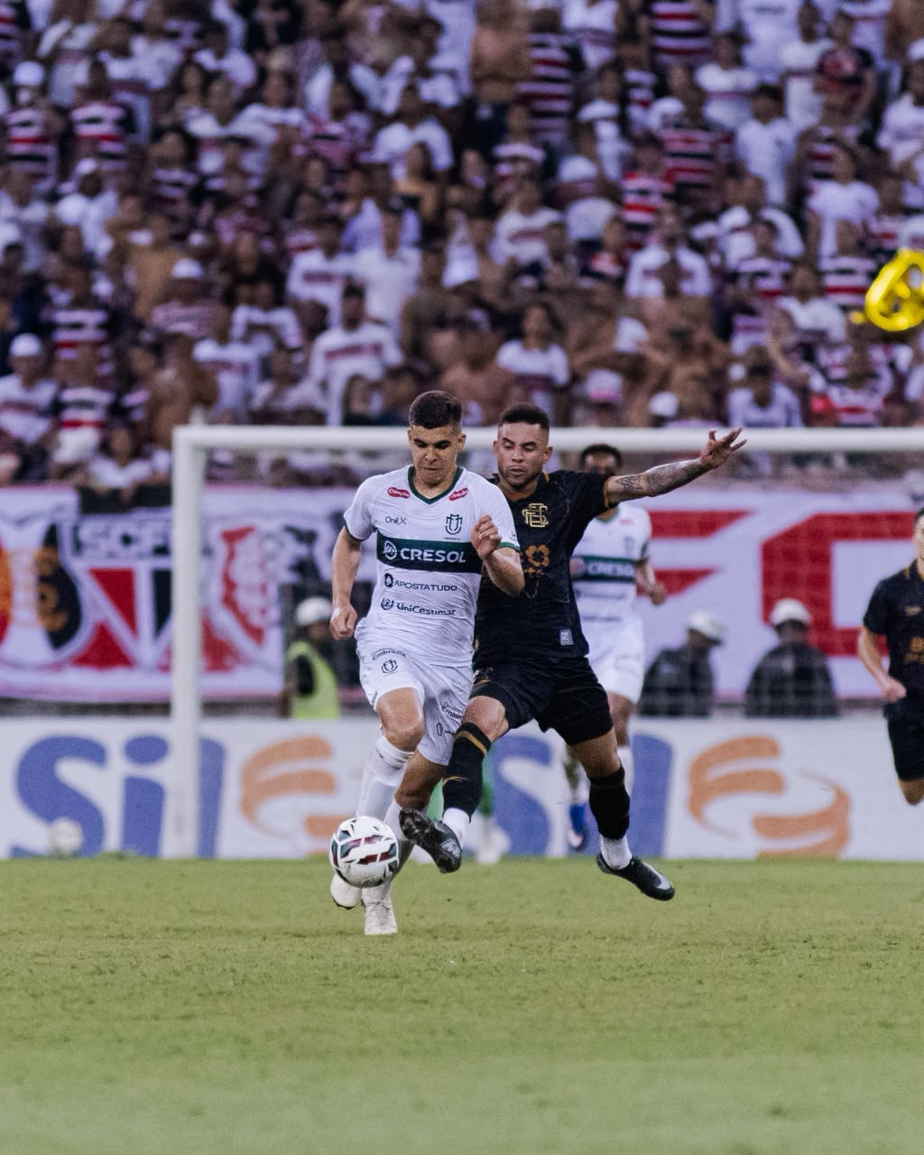 Jogador do Maringá conduz a bola diante de jogador do Santa Cruz, com a torcida ao fundo.