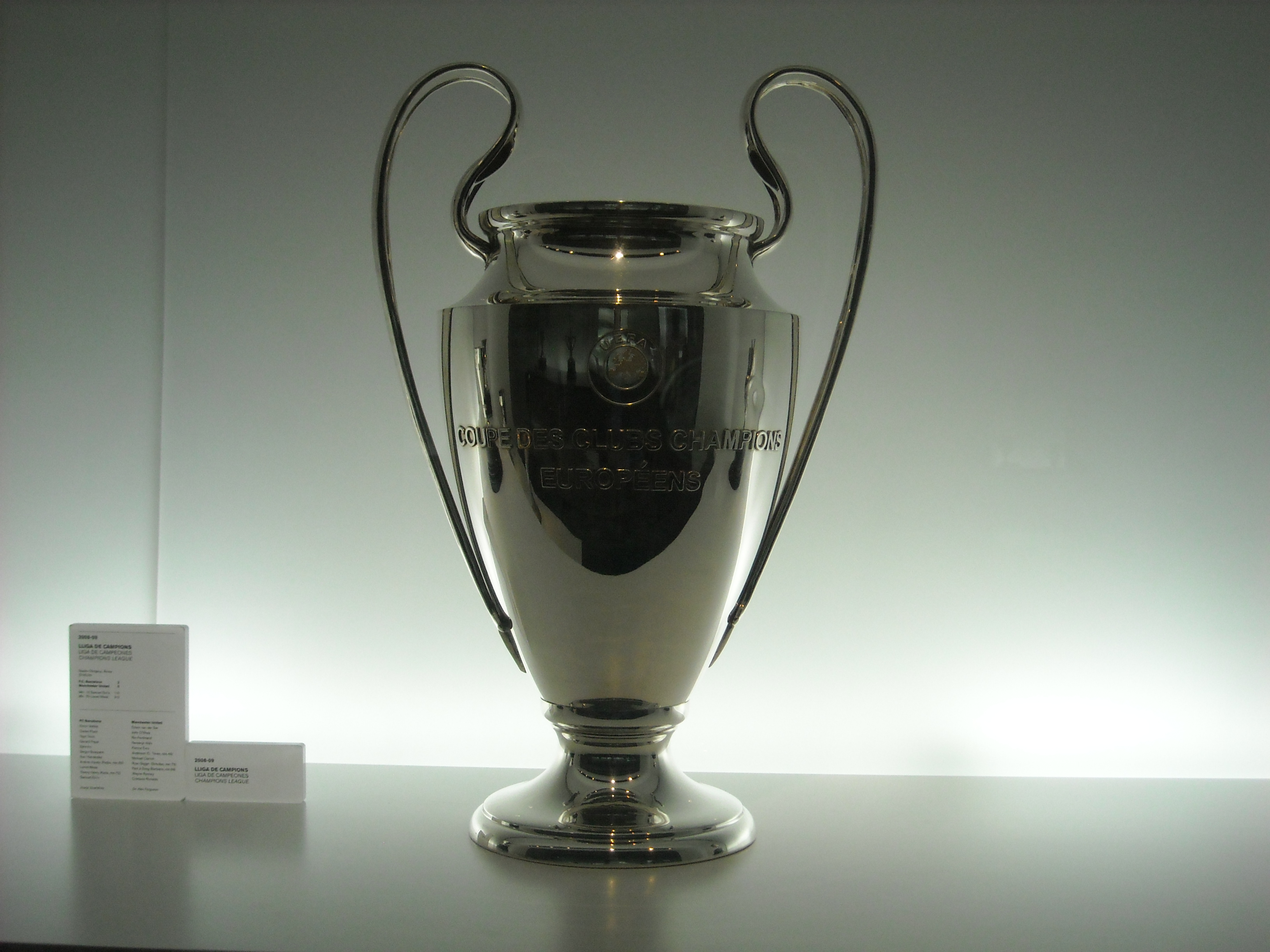 Taça prateada da UEFA Champions League em exposição, vista de frente sobre uma base.