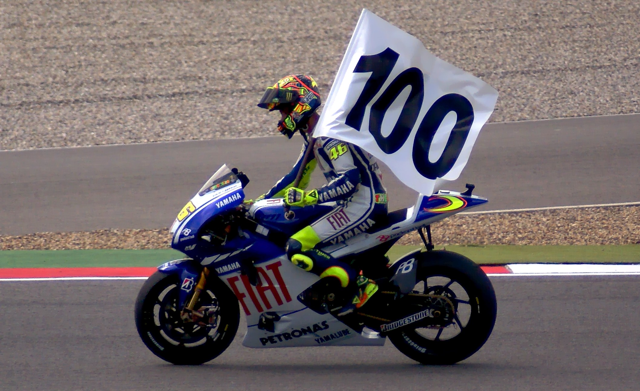 Valentino Rossi pilota a Yamaha número 46 carregando uma bandeira com o número 100.